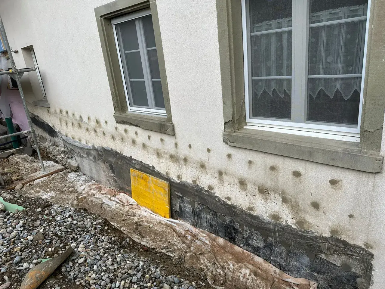 Arbeiten am Haus: Das Fundament trockenlegen mit neuer Abdichtung und Drainage. Gerüst und Baumaterialien sind sichtbar.