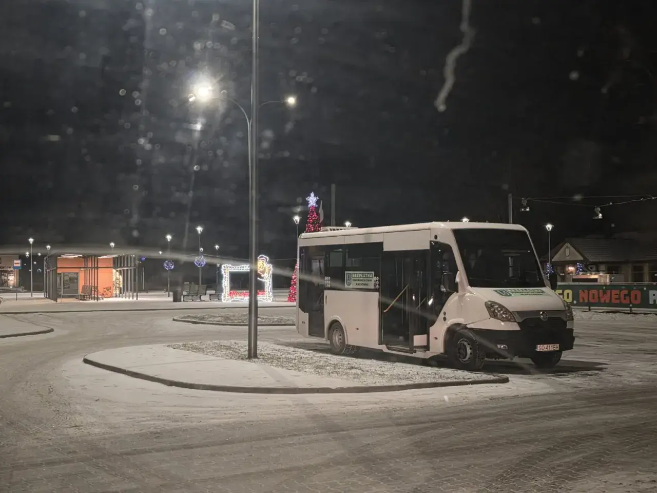 Biały bus na ośnieżonym przystanku nocą, ozdobionym świątecznymi lampkami.