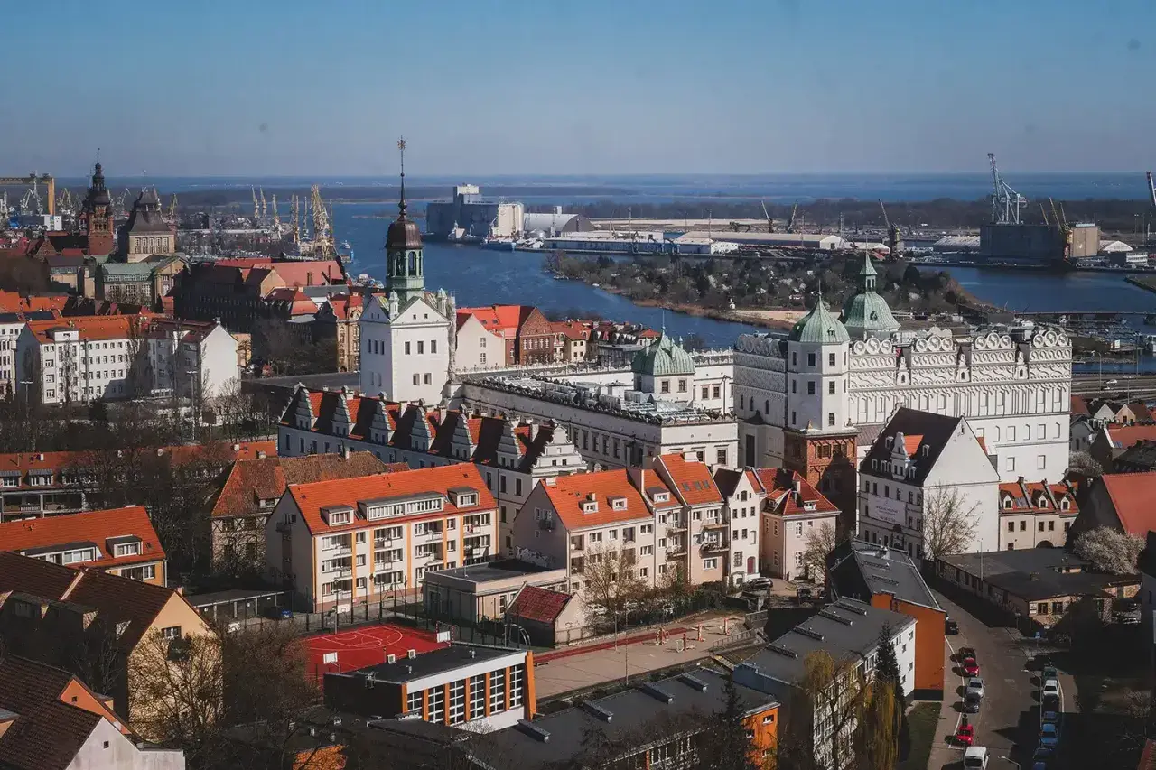 Widok na zamek i port w Szczecinie. Zabytkowe budowle z czerwonymi dachami kontrastują z błękitem wody i nieba.