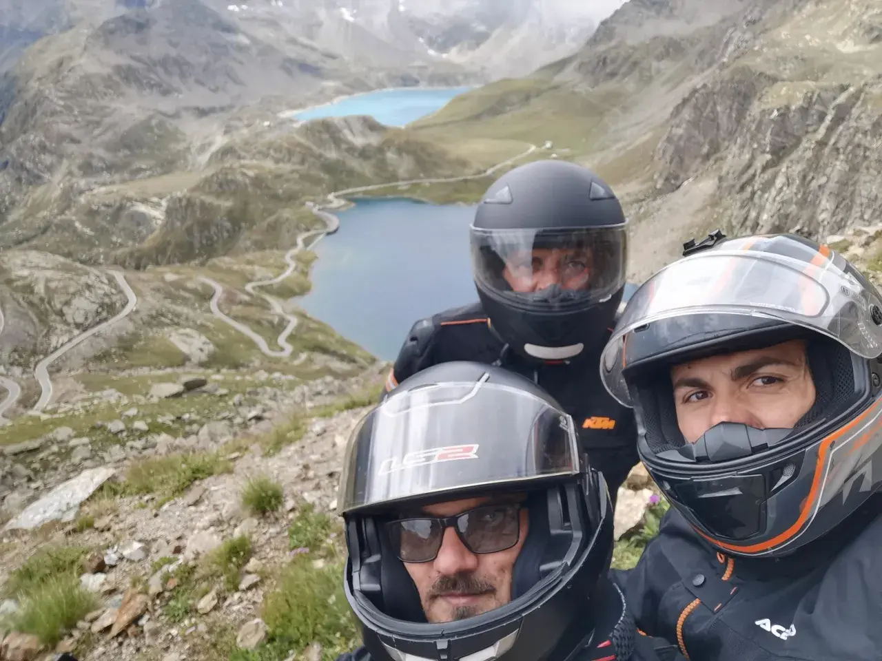 Tre motociclisti con caschi ammirano il panorama mozzafiato del Col del Nivolet, con i suoi laghi alpini e le strade tortuose.