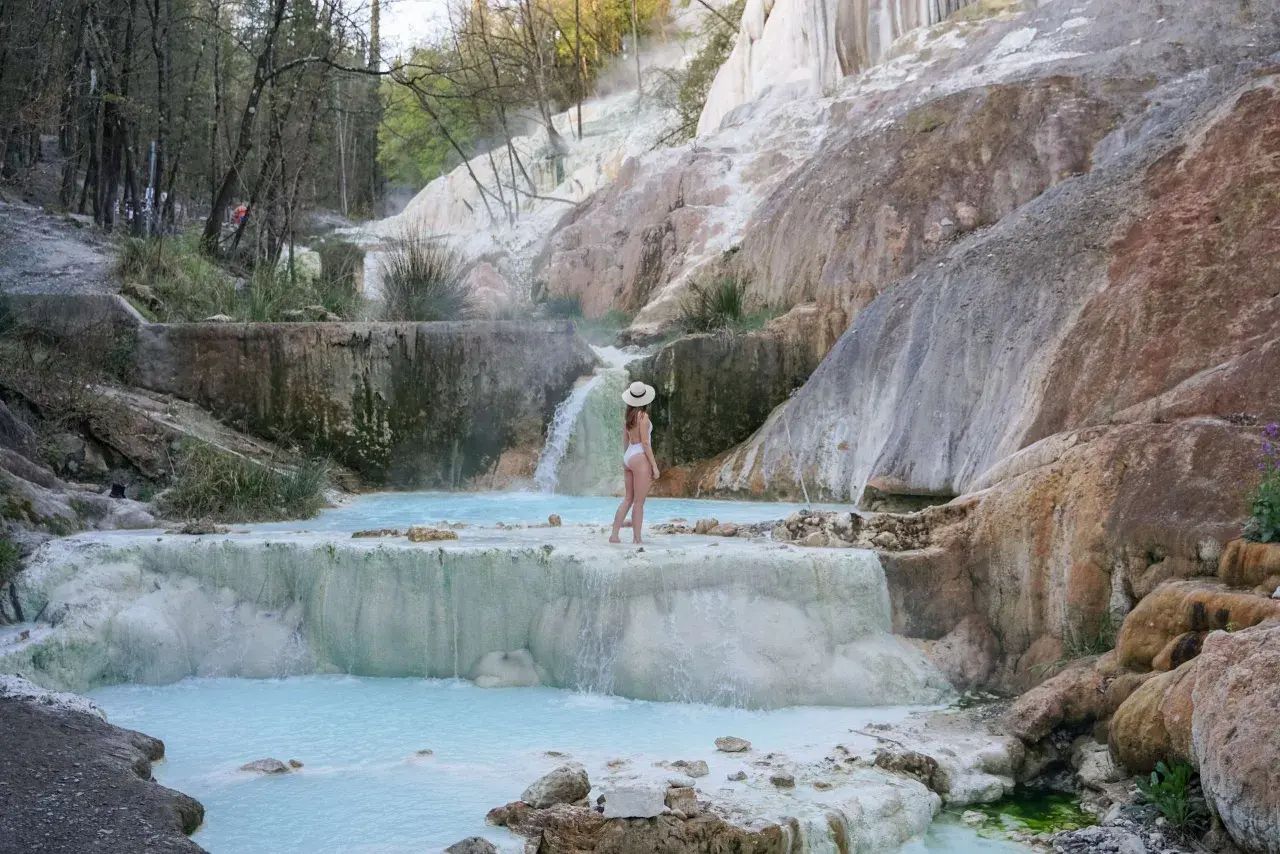 Une femme en maillot de bain blanc et chapeau de paille admire les cascades et les bassins turquoise des Bagni San Filippo, un paysage naturel spectaculaire.