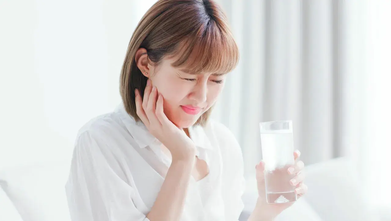 Donna con espressione sofferente, tiene un bicchiere d'acqua, soffre di ipersensibilità dentale.