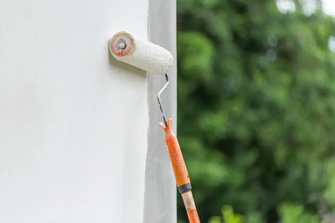 Fassade streichen ohne Reinigung: Ein Malerroller mit orangefarbenem Griff überzieht eine weiße Wand mit Farbe. Im Hintergrund verschwimmen grüne Bäume.