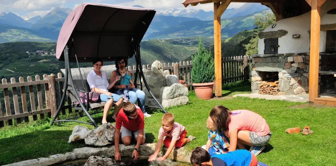 Famiglia rilassata in un giardino con vista sulle montagne, tipico delle fattorie altoatesine. Bambini giocano vicino a un laghetto, mentre adulti riposano su un'altalena.