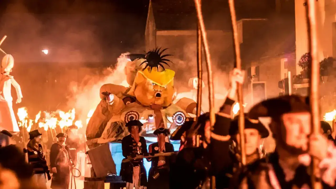 Un pupazzo gigante con un cappello giallo, circondato da fiamme e persone in costume, celebra un giorno festivo in inglese.
