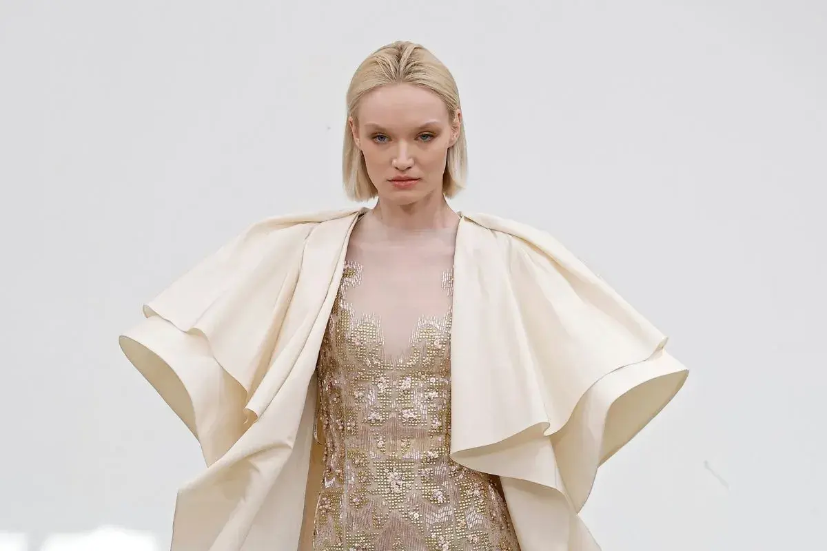 Modelka w kremowym, falbaniastym płaszczu i zdobionej sukni prezentuje dzieło haute couture.