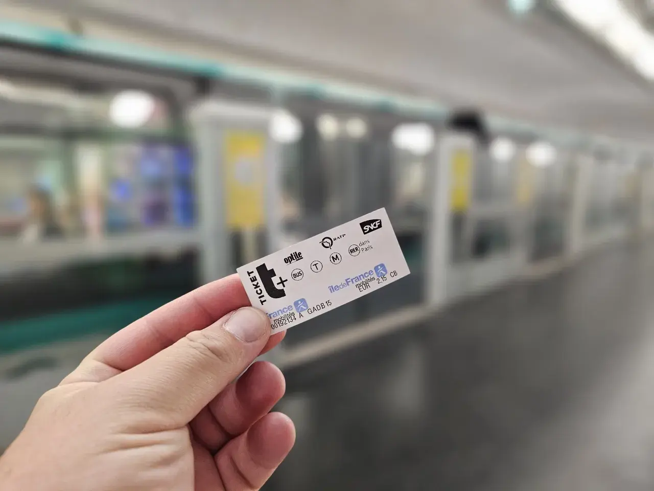 Dłoń trzyma bilet na metro paryskie. W tle widać pociąg metra.