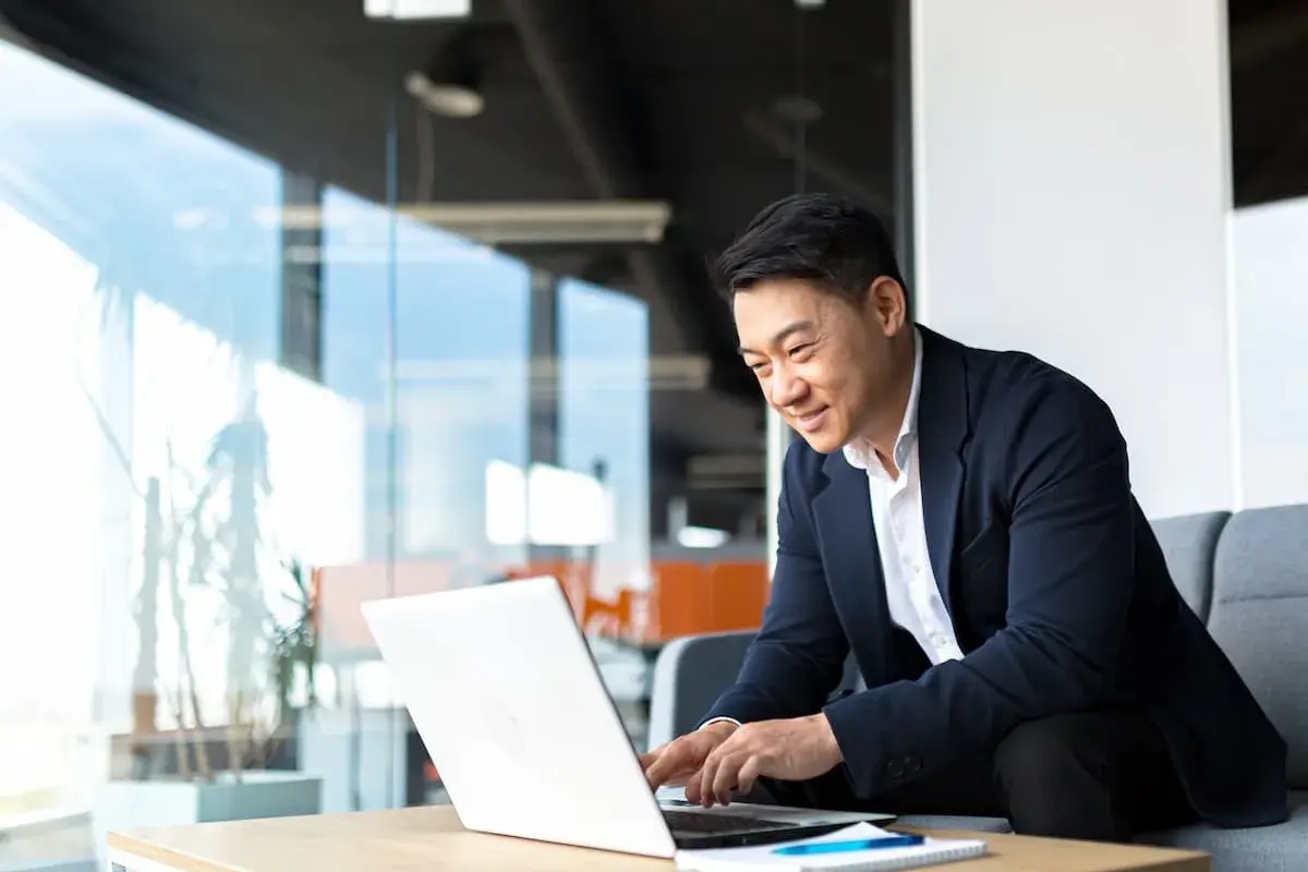 An Asian man in a suit smiles while working on his laptop, perhaps studying for his bachelors in organizational leadership.
