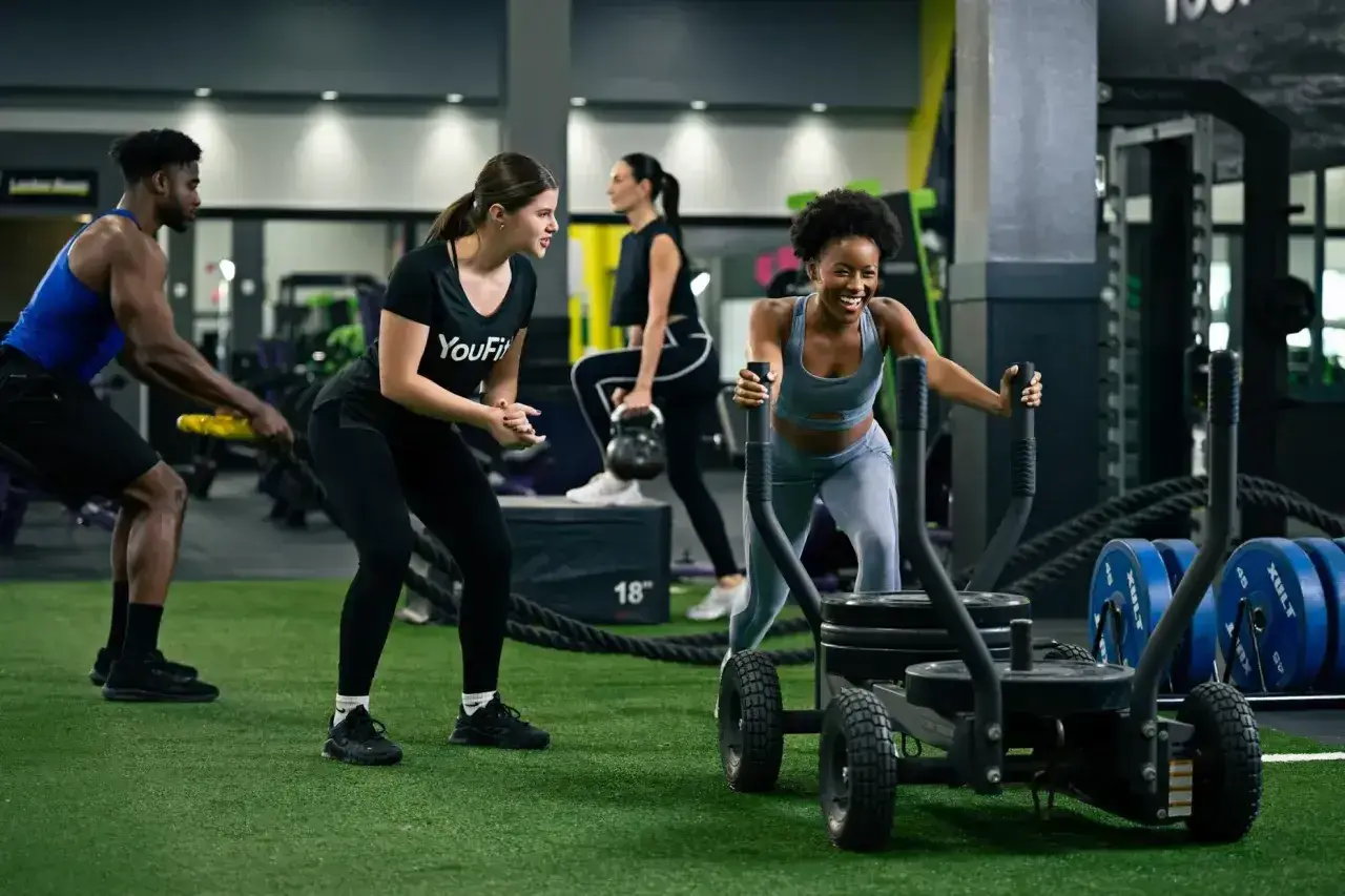 Intensywny trening interwałowy na siłowni. Kobieta pcha sanie, mężczyzna ćwiczy z linami, a inni wykonują ćwiczenia z kettlebell.