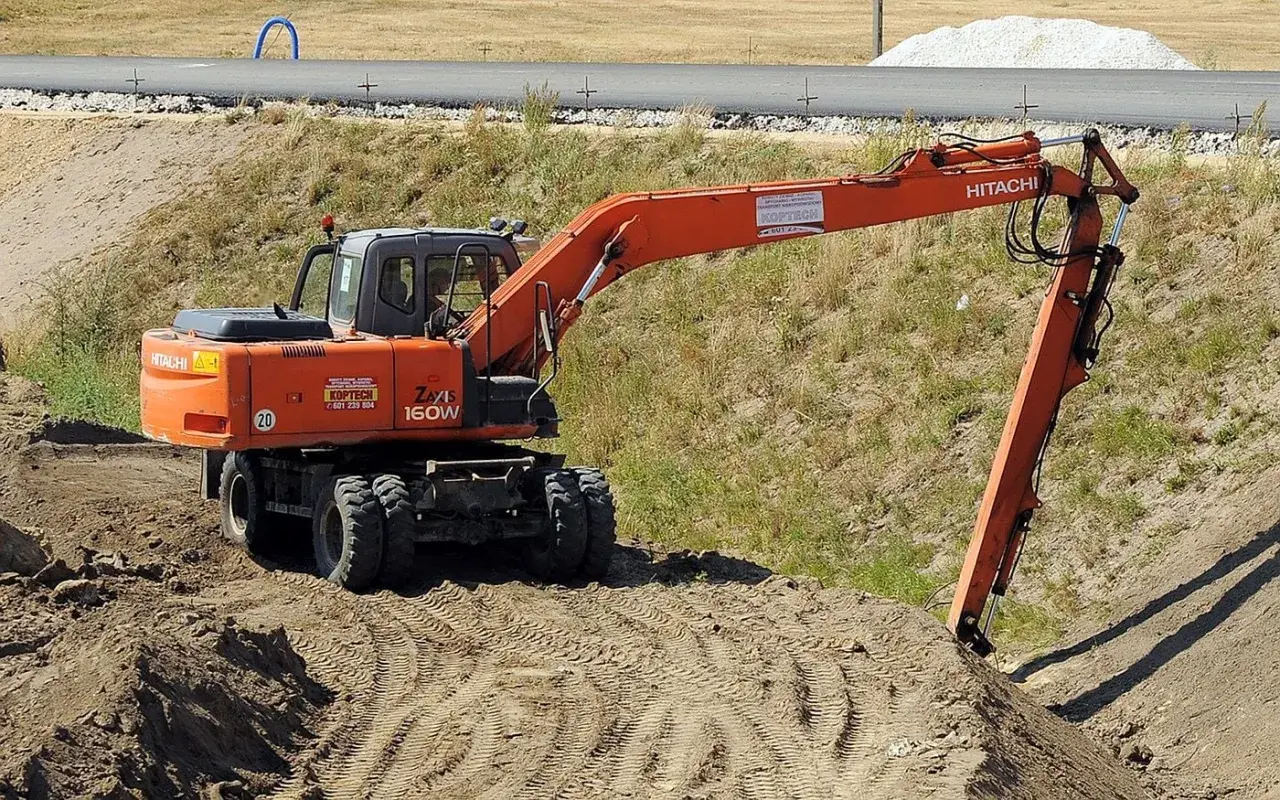 Pomarańczowa koparka Hitachi Zaxis 160W pracuje na budowie. Operator posiada odpowiednie uprawnienia na koparki, by sprawnie wykonywać prace ziemne.