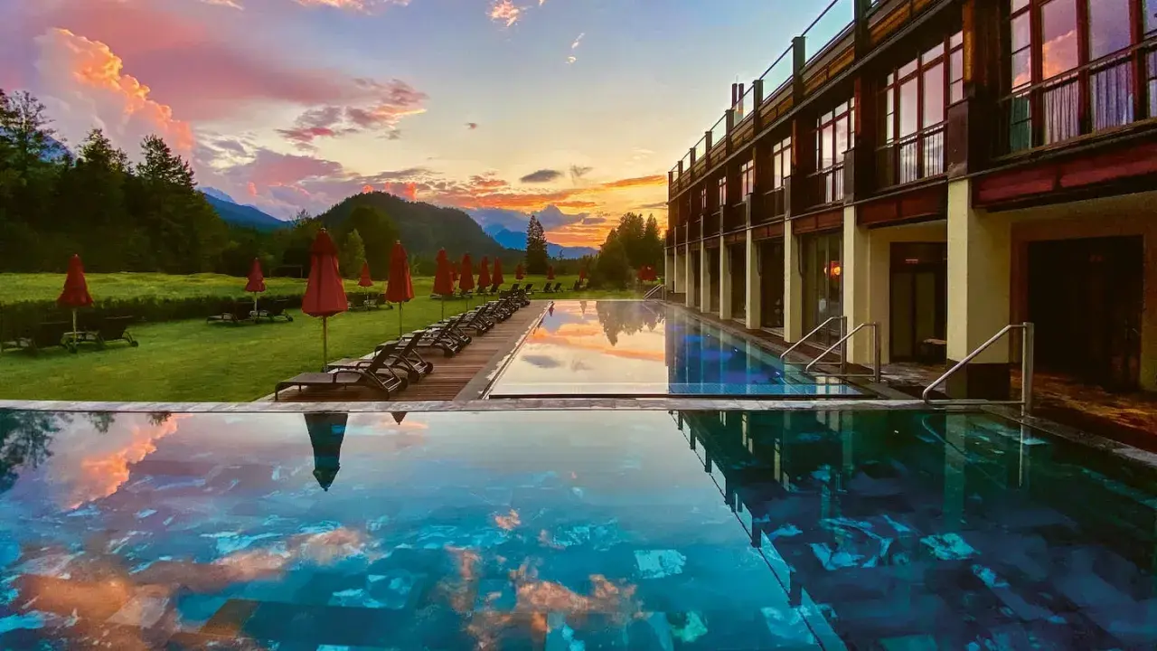Abendrot über den Alpen spiegelt sich im Infinity-Pool eines der besonderen Hotels Bayern. Liegestühle und rote Sonnenschirme säumen den Rasen.