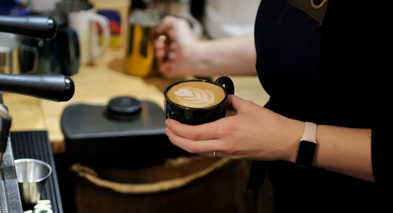 Barista trzyma kubek z flat white, ozdobiony latte art. Pyszna kawa, której kcal nie są wysokie.