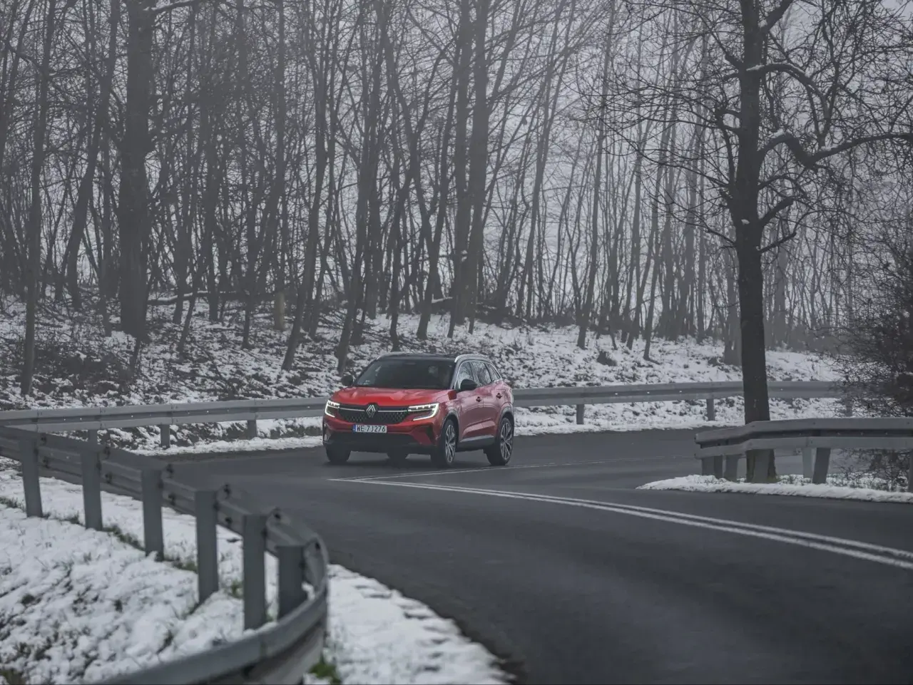 Czerwony SUV jedzie po mokrej drodze, otoczonej ośnieżonym lasem. Kierowca uważa, bo to może być gololedz.