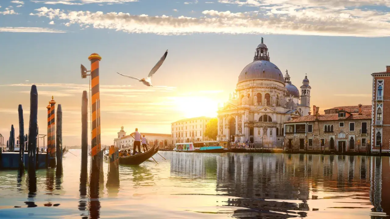 Góndola navega por canal veneciano al atardecer, con la Basílica de Santa Maria della Salute y otras **ciudades de Europa** reflejadas en el agua.
