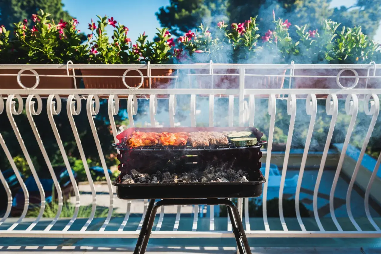 Grill z mięsem i warzywami na balkonie, w tle kwiaty i błękitne niebo.