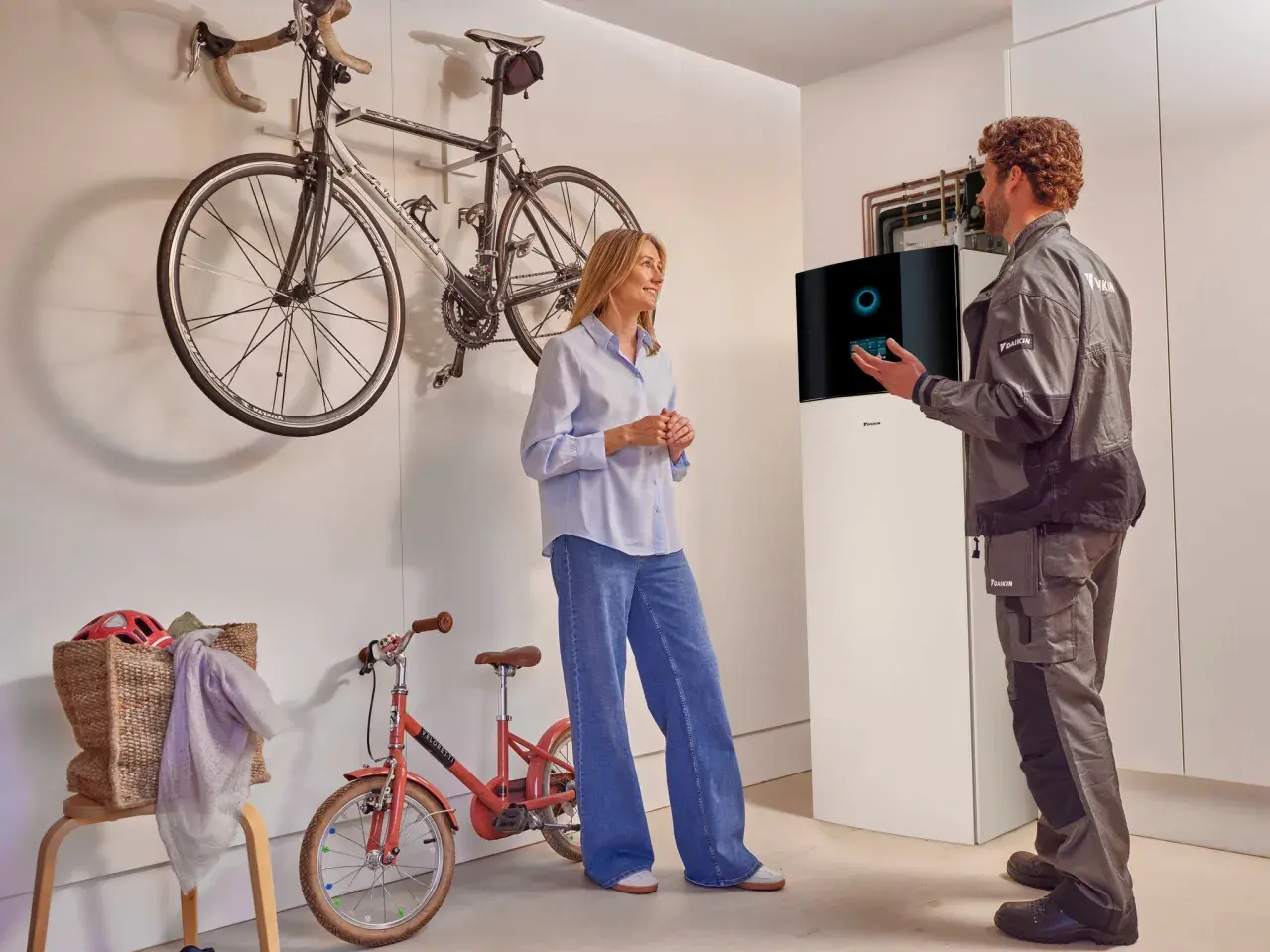 Técnico explica a una mujer el **rendimiento estacional aerotermia** junto a una unidad Daikin. Bicicletas colgadas al fondo.