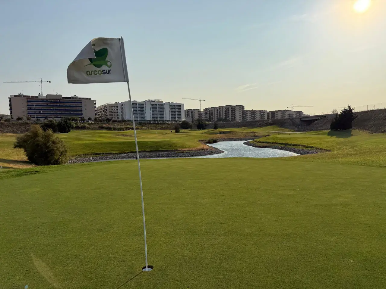 Bandera de Arcosur en un campo de golf, con edificios residenciales y grúas de construcción al fondo.