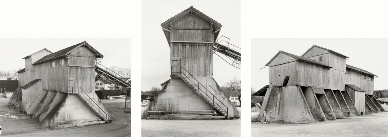 Strutture industriali in legno e cemento, tipiche delle opere di Hilla Becher, con scale esterne e nastri trasportatori.
