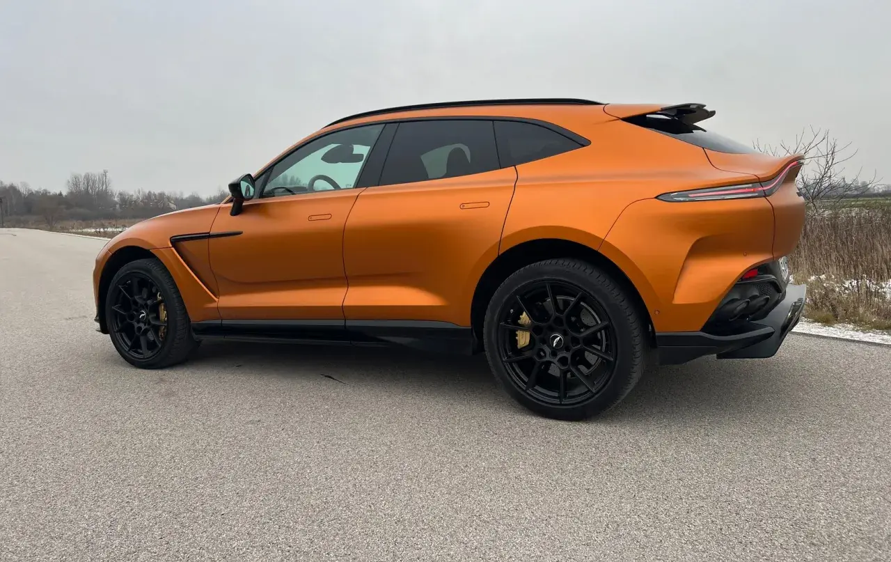 Pomarańczowy Aston Martin DBX z czarnymi felgami i przyciemnianymi szybami na tle pochmurnego nieba.