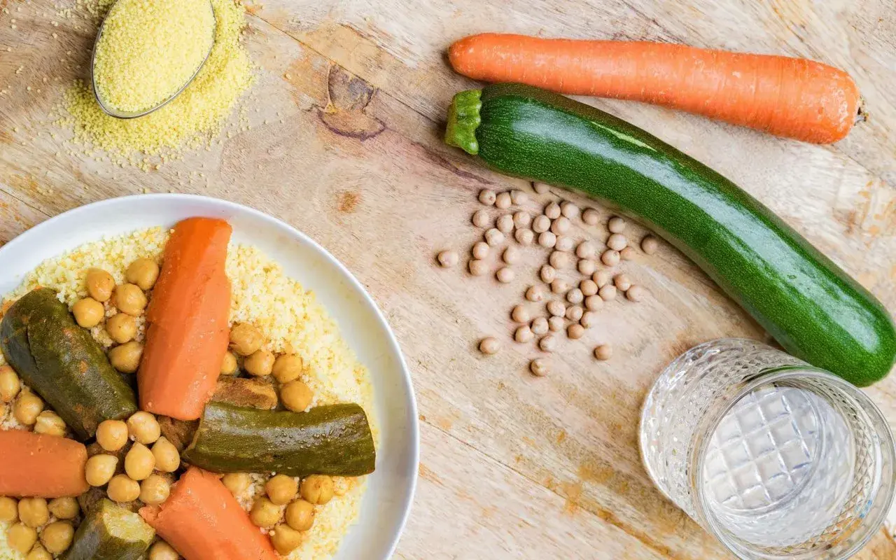 Un plat de couscous aux légumes, pois chiches, carottes et courgettes. Une cuillère de semoule est à côté.