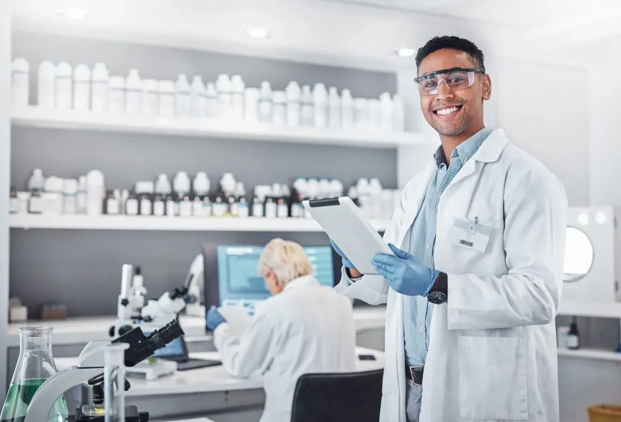 Uśmiechnięty technik farmacji w okularach ochronnych trzyma tablet w laboratorium, gdzie na półkach stoją liczne butelki, a w tle pracuje inna osoba.