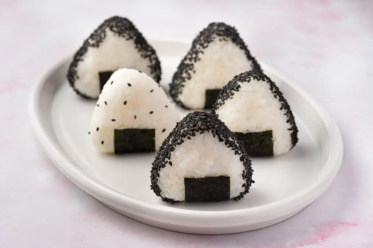Five triangular rice balls, some coated in black sesame seeds, sit on a white plate. This is what a rice ball looks like.