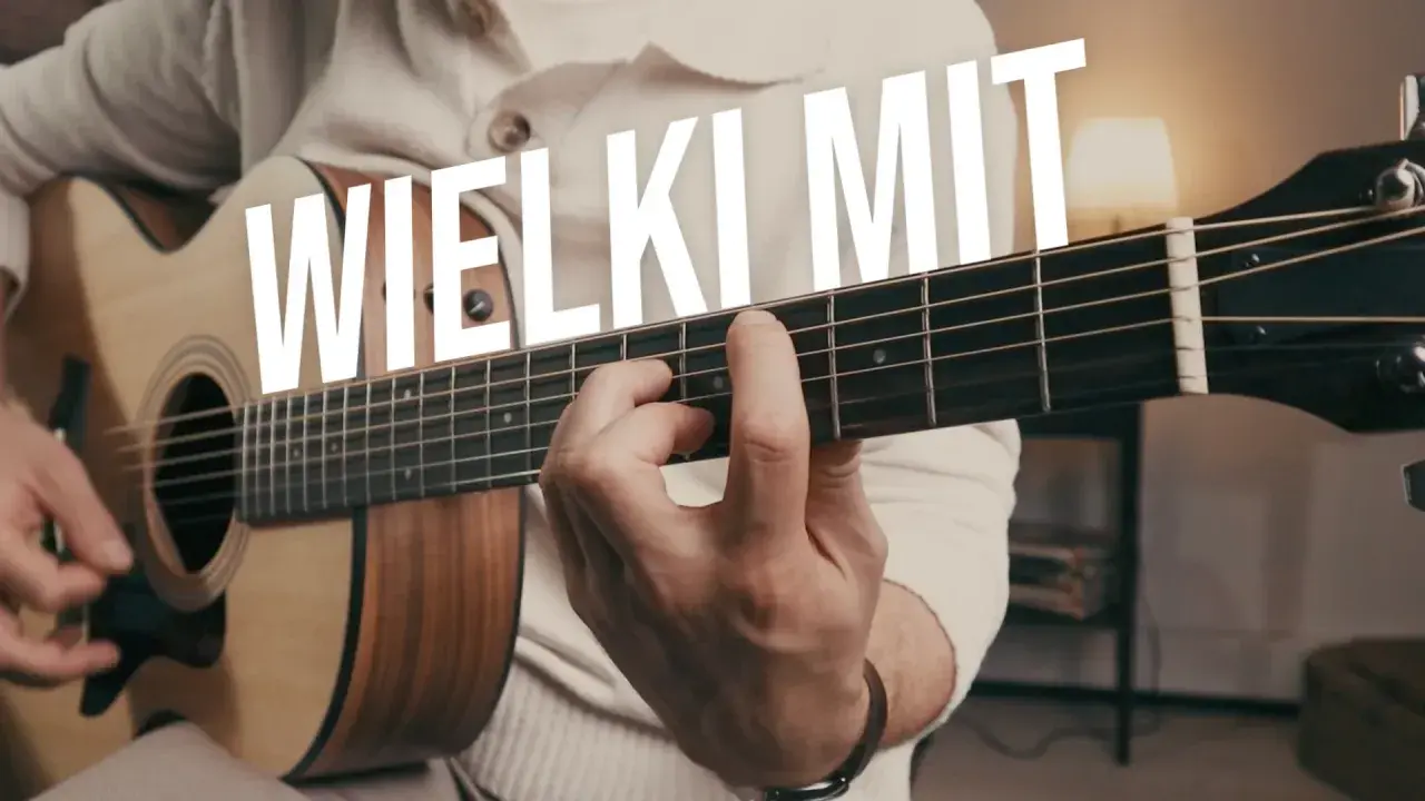 Ręka grająca na gitarze akustycznej, palce na gryfie. W tle biały napis "WIELKI MIT". Idealne ćwiczenia gitarowe.