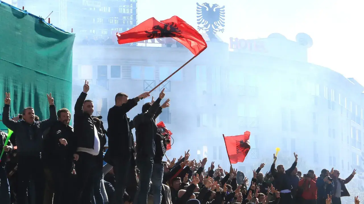 Människor viftar med albanska flaggor och gör fredstecken. En del av folkmassan verkar fira, vilket ger en känsla av stolthet och gemenskap. Detta kan vara en del av fakta om Albanien.