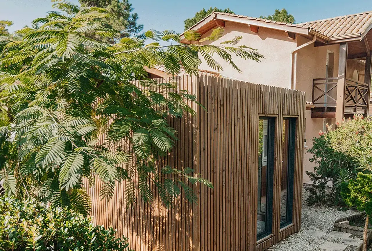 Extension de maison en bois moderne, bardage vertical, fenêtres sombres. Un espace de vie supplémentaire sans permis de construire, intégré dans un jardin verdoyant.
