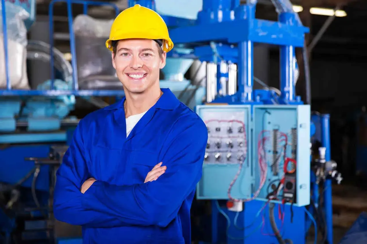 Uśmiechnięty pracownik w żółtym kasku i niebieskim kombinezonie, stojący przed panelem elektrycznym. To może być pracownik hurtowni elektrotechnicznej Comel Rumia.