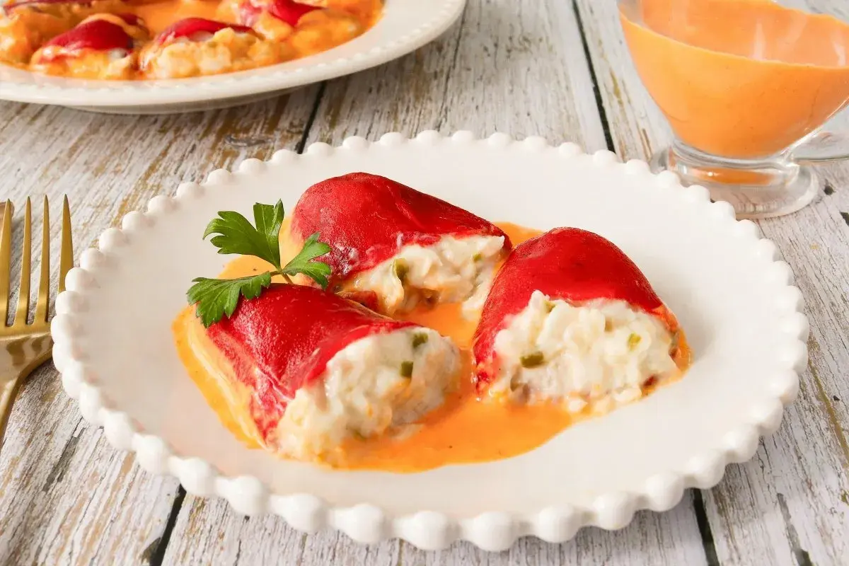 Pimientos rellenos de bacalao, cubiertos en una cremosa salsa naranja, servidos en un plato blanco con un tenedor dorado al lado.