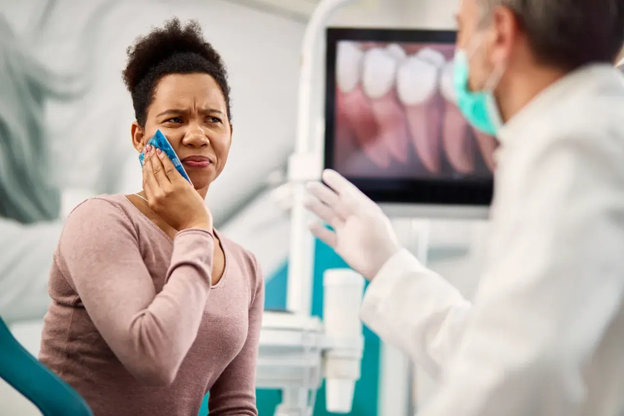 Mujer con compresa fría en la mejilla, sufriendo un alveolitis con dolor insoportable, mientras el dentista le explica algo.