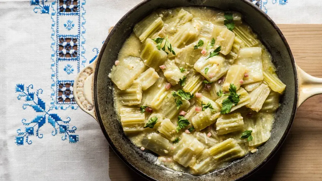 Cardo en salsa cremosa con trocitos de jamón y perejil, servido en sartén sobre mantel bordado. Un plato delicioso con cardo y almendras.