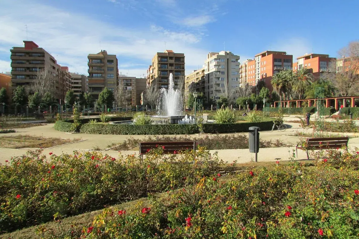 Parque urbano con fuente central y edificios residenciales al fondo. Los viveros de Valencia lucen sus rosales en flor.