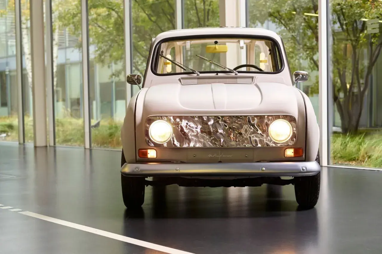 En ljusbeige Renault 16 står framför stora fönster med grönska utanför. Bilens front med sina runda strålkastare och kromade grill är i fokus.