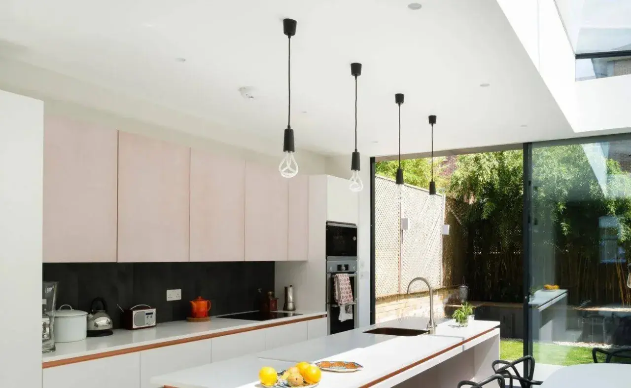 Cocina moderna con gabinetes rosados y una lámpara de barra colgante sobre la isla. Ventanales dan a un jardín exuberante.