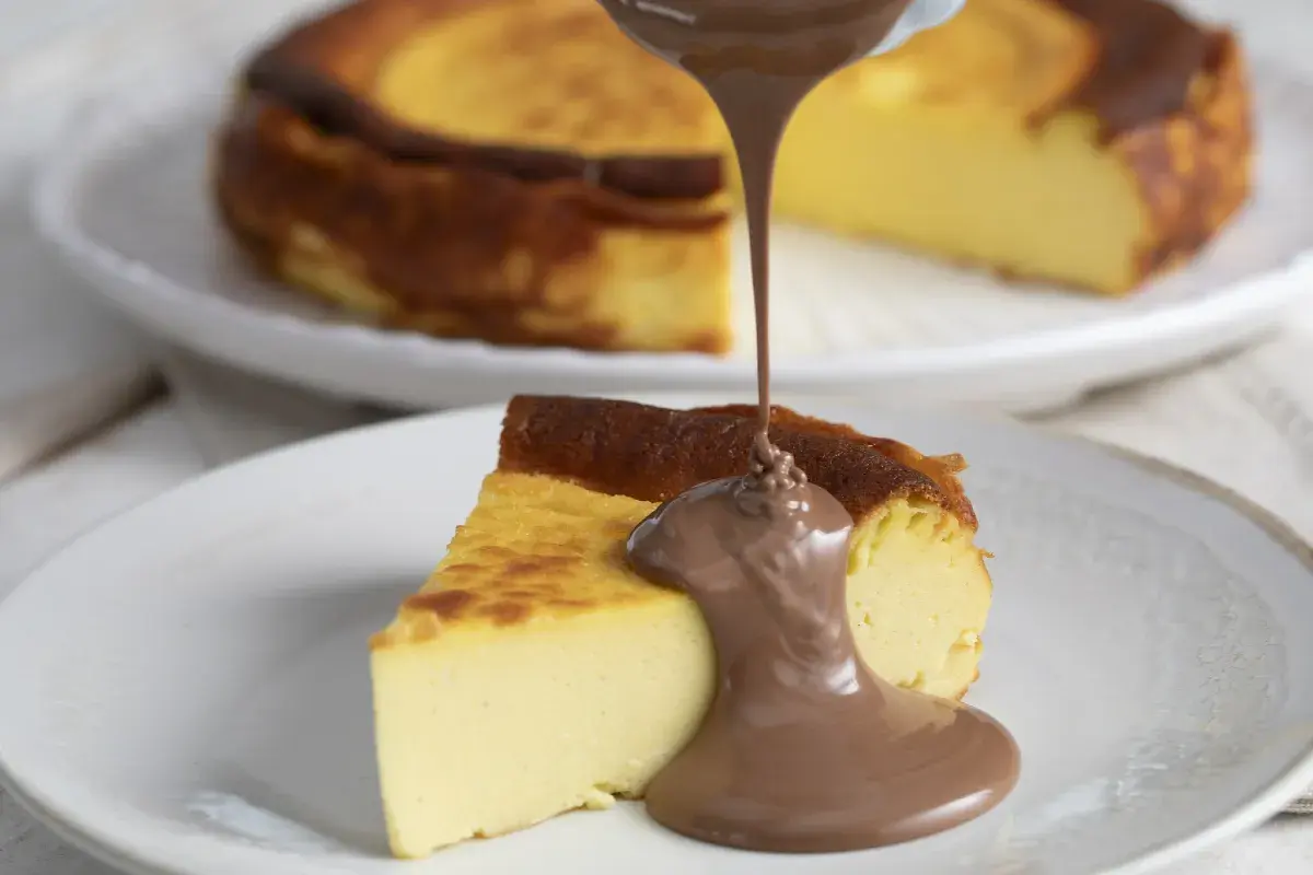 Un trozo de basque cheesecake cremoso recibe un chorro de chocolate fundido. Otro pastel entero se ve desenfocado al fondo.