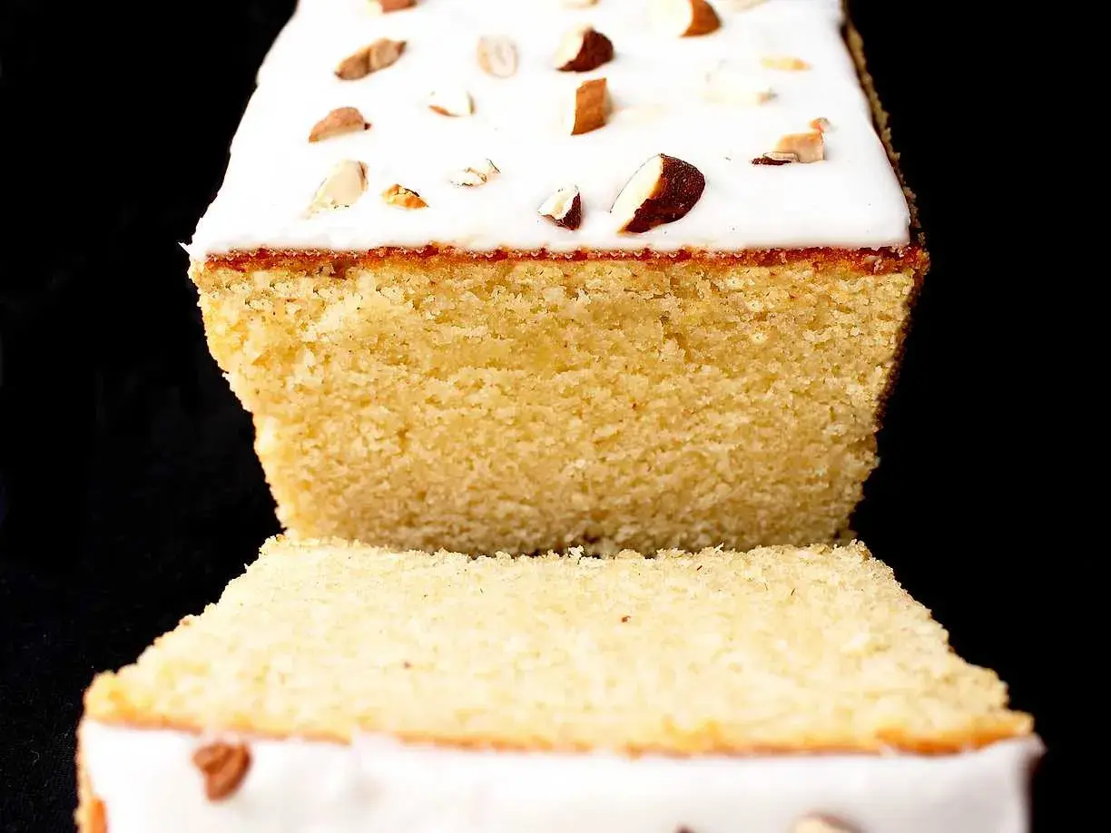 A slice of almond cake with white icing and chopped almonds on top, against a black background.