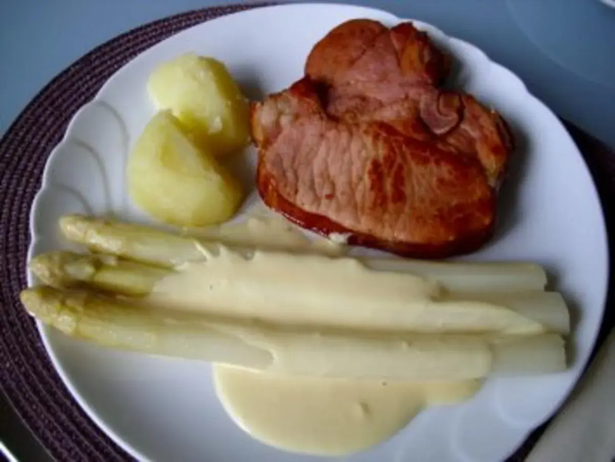 Ein Teller mit gebratenem Schinken, Kartoffeln und Spargel in Sauce Hollandaise. Der Spargel ist in Alufolie gewickelt.