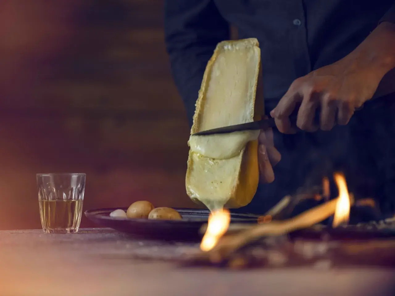Un morceau de fromage à raclette fond sur un feu, rappelant son origine alpine. Des pommes de terre et un verre de vin complètent la scène.