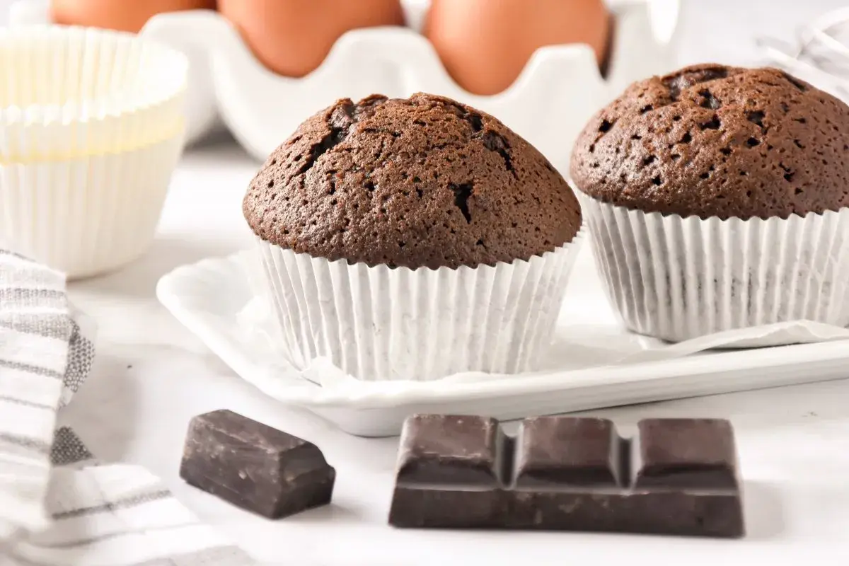 Dos magdalenas de chocolate recién horneadas, listas para disfrutar. A su lado, trozos de chocolate y moldes para hornear.