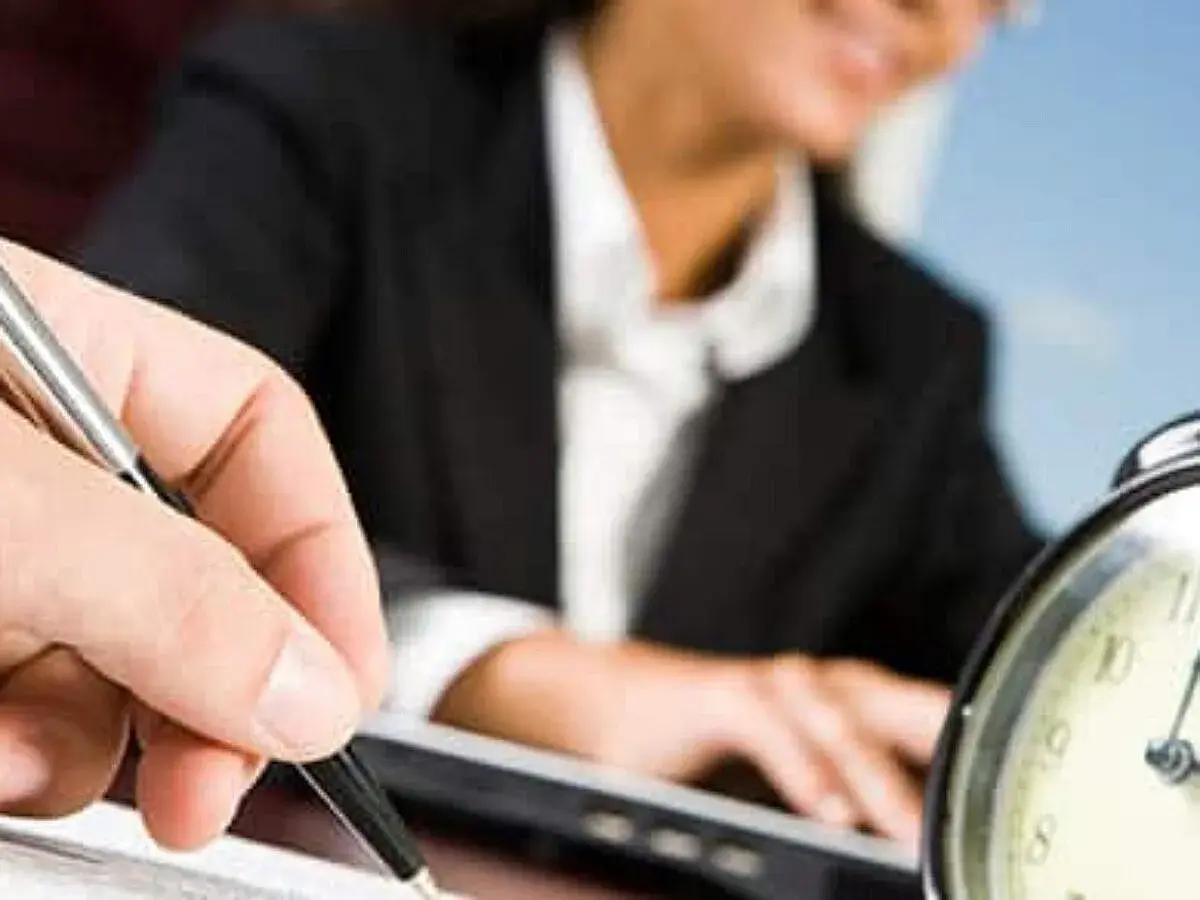A hand holds a pen, poised to take notes, as a person in a suit works at a laptop. This scene evokes the process of creating board meeting minutes examples.