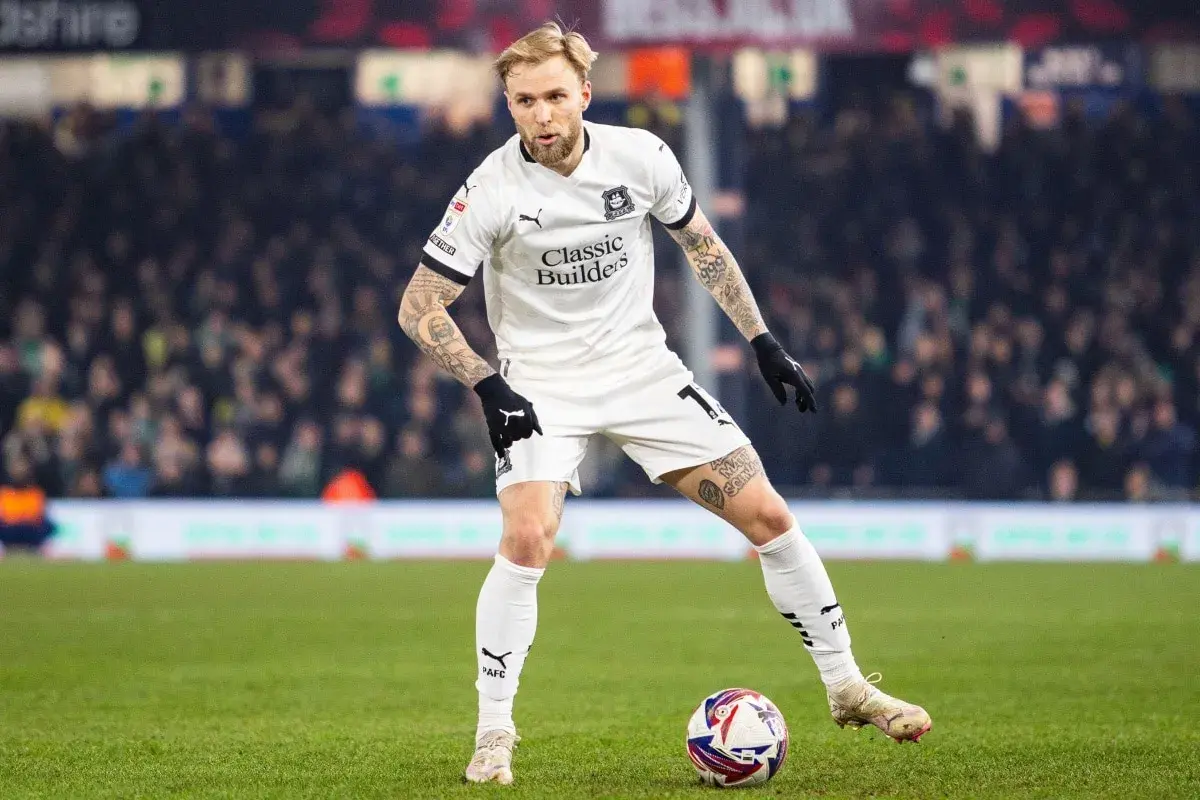 Piłkarz w białej koszulce z tatuażami na rękach i nogach, gdzie gra puchacz, przygotowuje się do strzału na zielonej murawie stadionu.