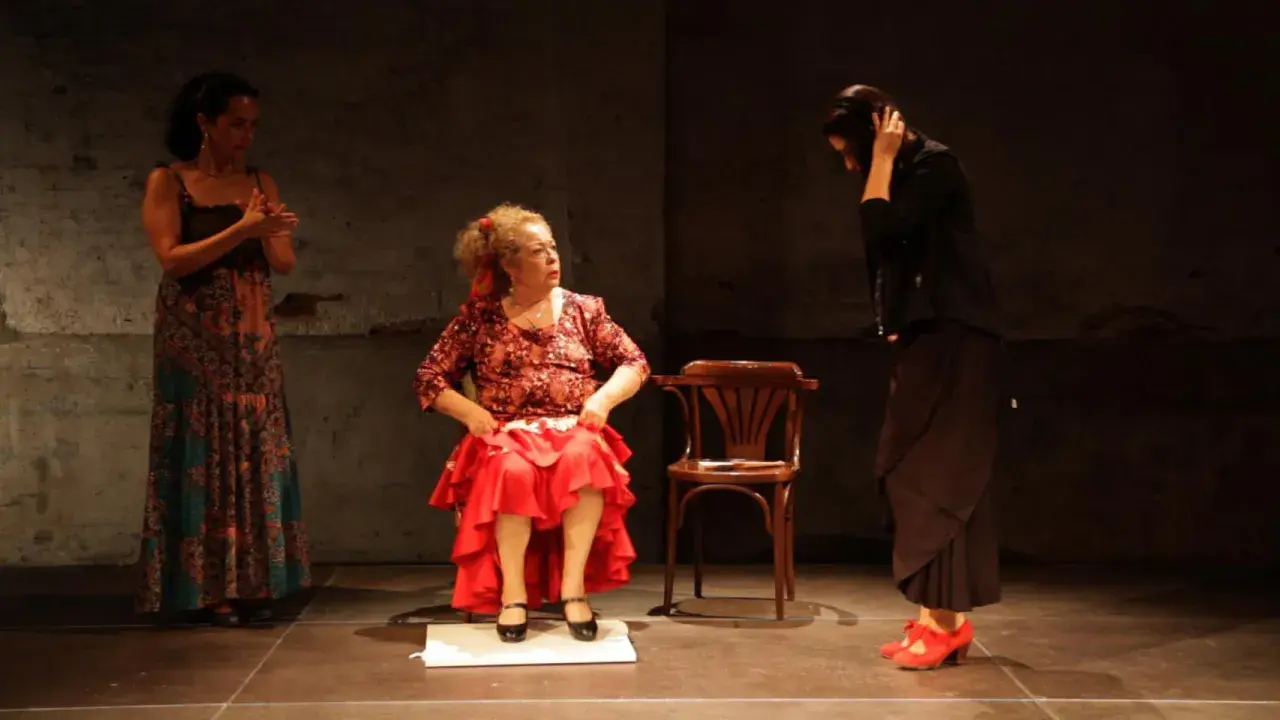 Tres mujeres en escena. Una sentada, con falda roja y blusa estampada, como **la Chana**. Otra de pie, con vestido largo y estampado. La tercera, de negro, con el pelo recogido.