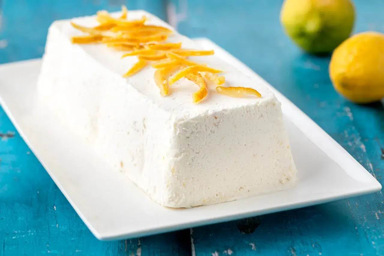Delizioso semifreddo al limone, guarnito con scorzette candite, su un piatto bianco. Due limoni freschi sullo sfondo.