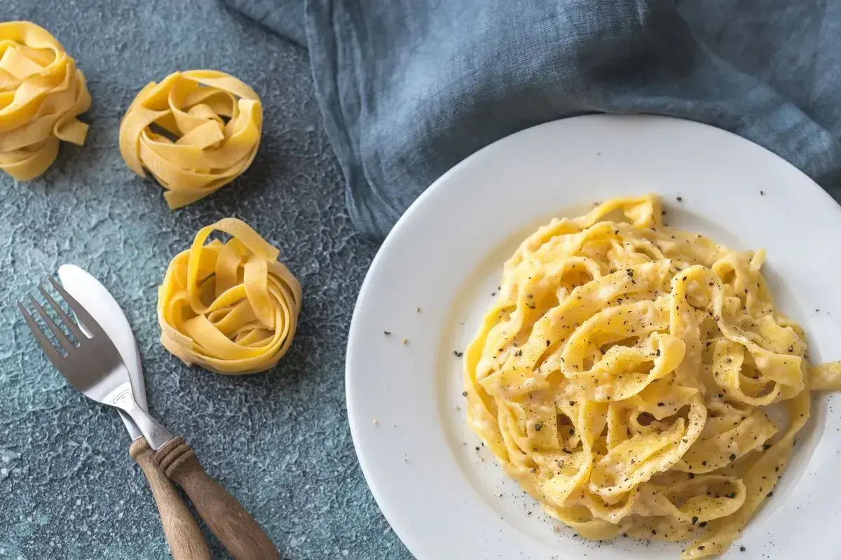 Un piatto di fettuccine alfredo cremoso, con nidi di pasta cruda e posate sul lato.