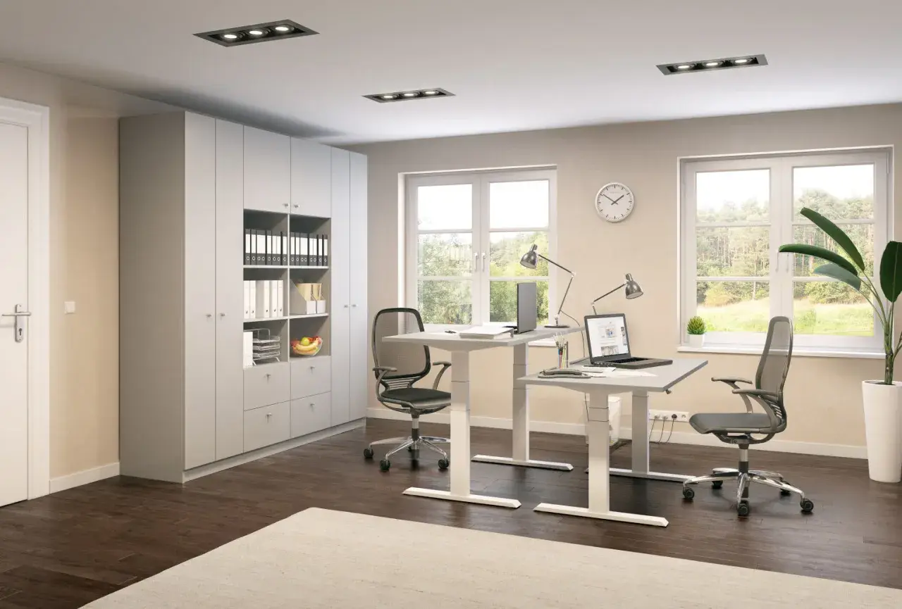 Ein helles, modernes Homeoffice mit zwei höhenverstellbaren Schreibtischen, Bürostühlen, einem großen Schrank und Fenstern mit Blick ins Grüne.