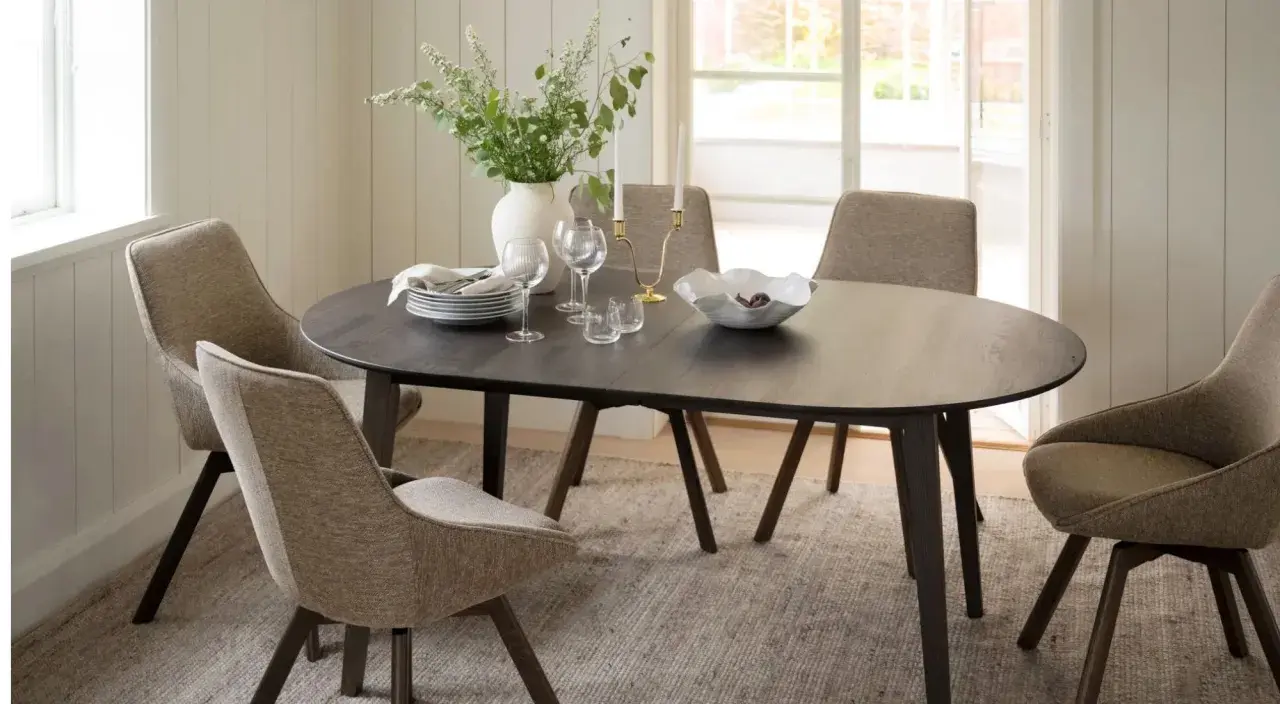 Ett runt mörkt matbord med fyra stoppade stolar runt sig. En vas med blommor, ljusstakar och en skål med frukt på bordet. En tidlös design klassiker.
