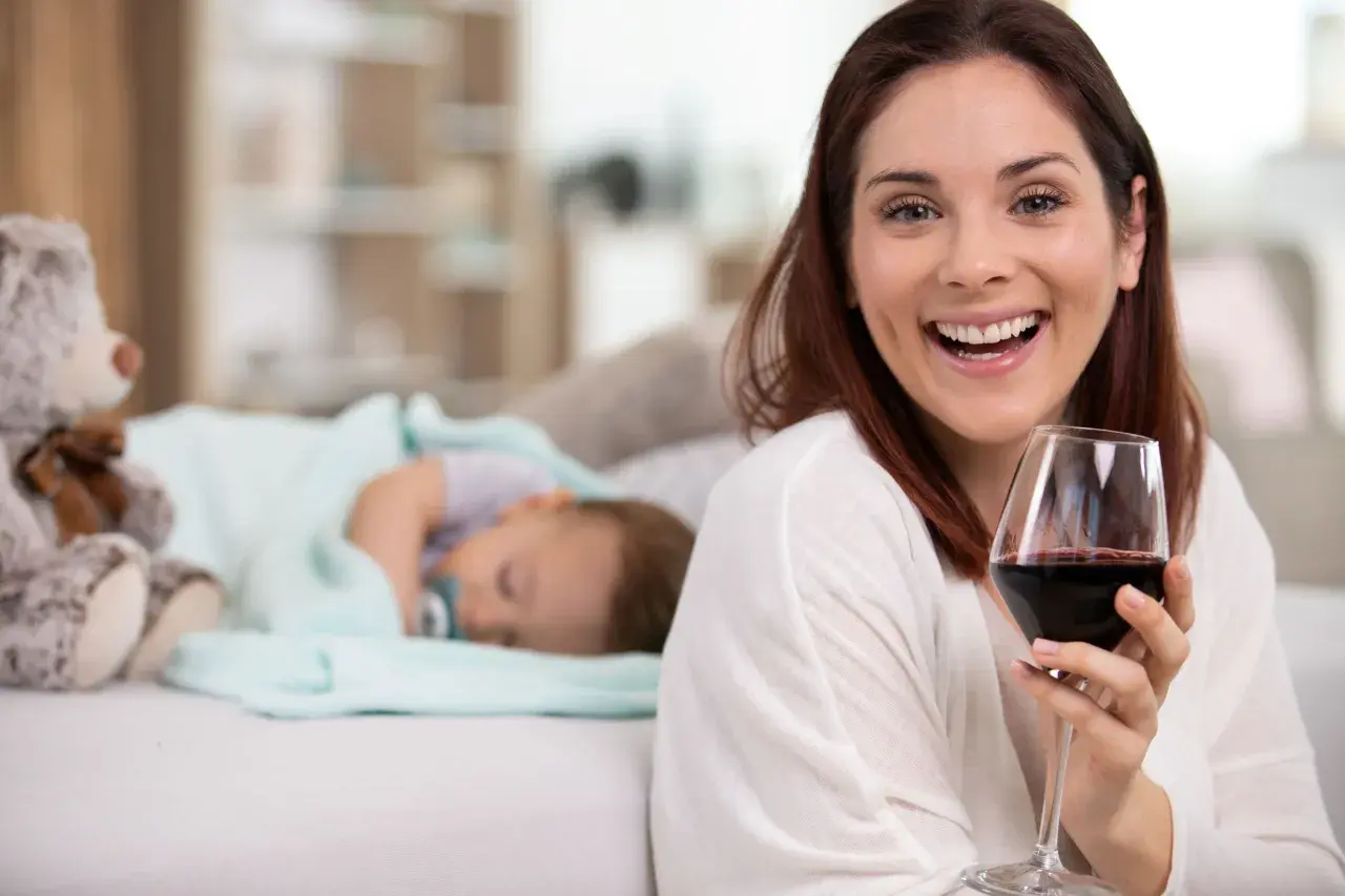 Uśmiechnięta kobieta z kieliszkiem czerwonego wina obok śpiącego dziecka. Czy można pić alkohol przy kamicy żółciowej?