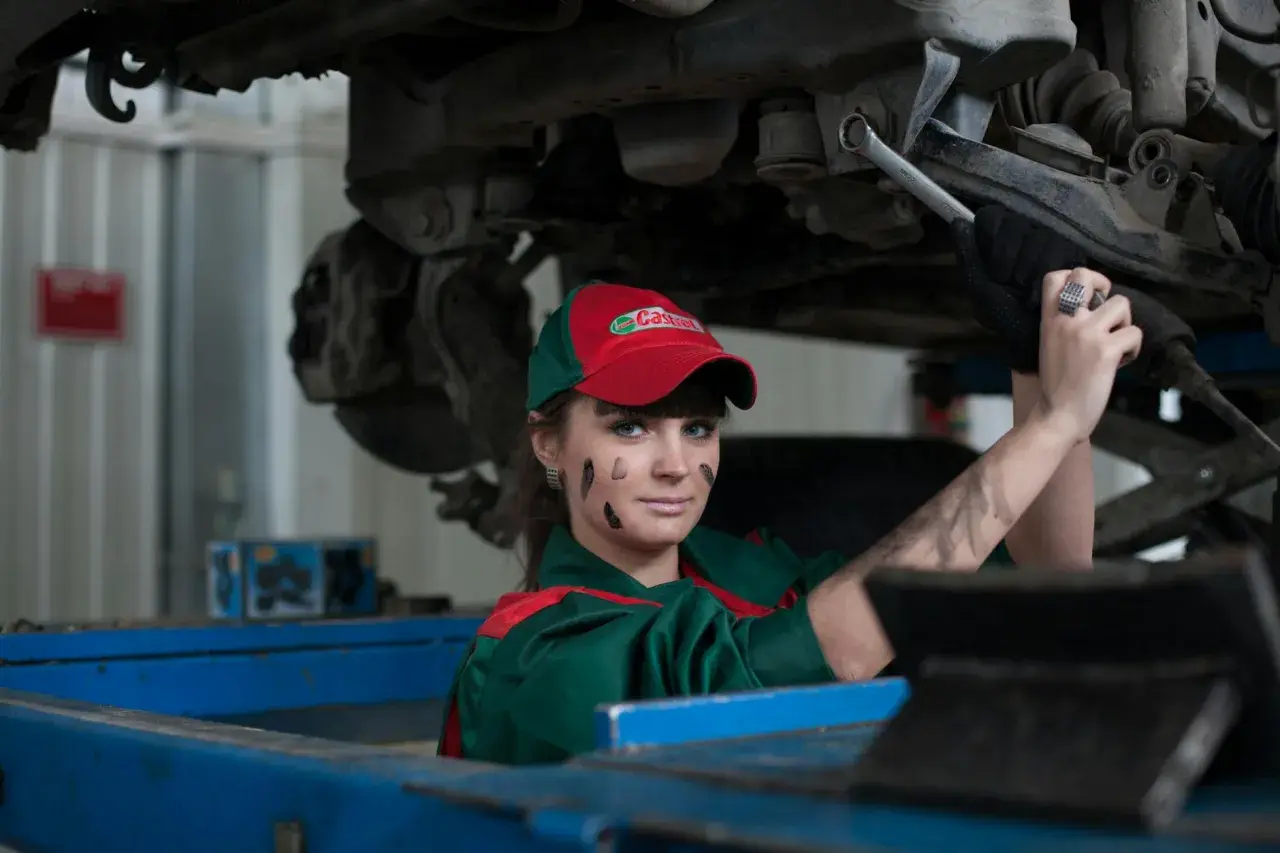 Mécanicienne sous une voiture, le visage barbouillé de cambouis. Elle est experte automobile, mais son regard suggère un abus de pouvoir.